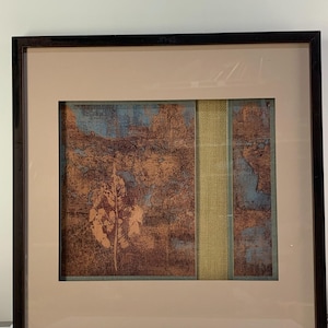 Könnte beinhalten: Gerahmter abstrakter Kunstdruck mit einem braunen Holzrahmen. Das Kunstwerk zeigt ein strukturiertes Design mit braunen, blauen und beigen Farben. Ein vertikaler Streifen aus hellgrünem Stoff teilt das Kunstwerk. Der Druck ist auf einem beigen Passepartout.