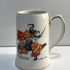 May include: Cream-coloured ceramic tankard with a gold rim and handle. It features a detailed illustration of a knight on horseback, with text reading "Thomas Beauchamp Earl of Warwick" and dates. A decorative item.