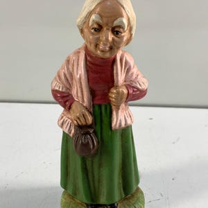 May include: A ceramic figurine depicting an elderly woman. She is dressed in a red top, a pink shawl, and a long green skirt. The figurine holds a small brown purse and stands on a grassy base. Her hair is white, and her face has painted details.