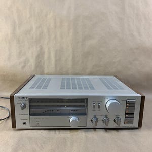 Puede incluir: Un receptor Sony STR-V35 vintage, plateado y con paneles de madera. La parte frontal presenta una pantalla de sintonizador, perillas de volumen y balance, y varios botones de función. La parte superior tiene un diseño ventilado.