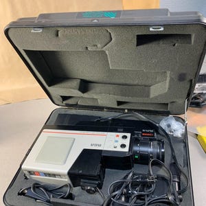 May include: Vintage RCA video projector in a black carrying case. The projector is white and black with red and blue accents. The case is open, revealing the projector, cables, and foam padding. The case has the text "VESTRO RACING" on the top.