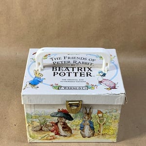 Peut inclure: Une boîte blanche avec une poignée, ornée d'illustrations des personnages de Pierre Lapin. La boîte a un fermoir doré et le texte "THE FRIENDS OF PETER RABBIT BY BEATRIX POTTER" imprimé sur le devant.
