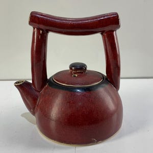 May include: A maroon ceramic teapot with a distinctive arched handle. The teapot has a rounded body, a spout, and a lid with a small knob. The rim of the lid has a black accent. The teapot is designed for brewing tea.