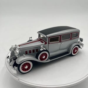May include: A silver classic car model with a black roof and red accents. The car has red wheels with white rims and chrome detailing. The model is a vintage style car.