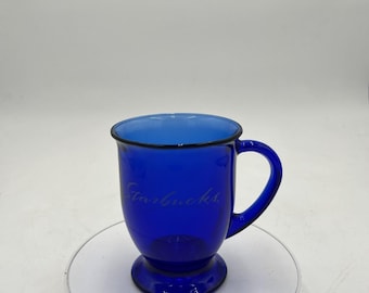 Puede incluir: Una taza de café Starbucks azul cobalto con borde y asa negros. El logotipo de Starbucks está grabado en el lateral del cristal. La taza tiene una base de pedestal y está sobre una superficie blanca.