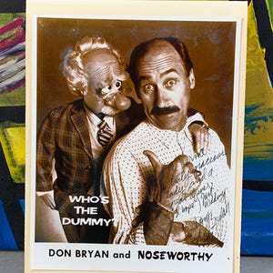May include: Sepia-toned photograph of Don Bryan and Noseworthy, a ventriloquist and his dummy. The image includes the text "WHO'S THE DUMMY?" and the names "DON BRYAN and NOSEWORTHY". The dummy has a large nose and is wearing glasses.