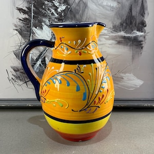 May include: A decorative ceramic pitcher with a yellow body, blue handle, and a blue rim. The pitcher features colorful floral and swirl designs in blue, green, and red. The base has a red, yellow, and black stripe.
