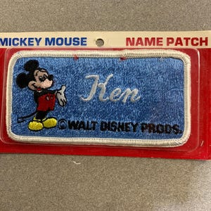 Könnte beinhalten: Blauer Mickey Mouse Namensaufnäher mit dem Namen "Ken" in Weiß gestickt. Der Aufnäher zeigt Mickey Mouse und den Text "WALT DISNEY PRODS." sowie "NAME PATCH".