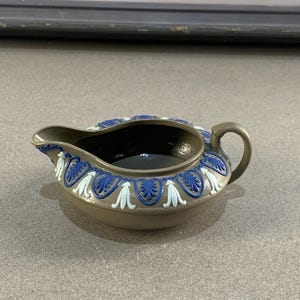 Puede incluir: Una pequeña jarra de crema marrón con asa y boquilla. El borde está decorado con diseños florales azules y blancos. El interior es oscuro. La jarra de crema está sobre una superficie clara.