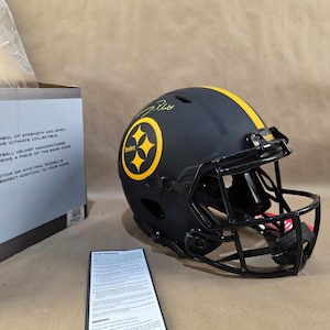 Peut inclure: Un casque de football américain noir avec une bande jaune et le logo des Steelers de Pittsburgh. Le casque a un masque noir et une signature sur le dessus. Une feuille d'avertissement et une boîte sont à côté du casque.