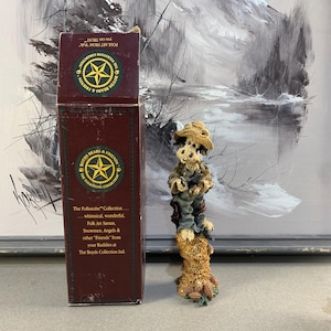 May include: A Boyd's Bears & Friends figurine of a bear wearing a hat and overalls, standing on a stack of corn. The figurine is next to a maroon box with text that reads "The Folkstone Collection". The background is a painting of a winter landscape.