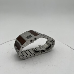 May include: A silver wristwatch with a rectangular face and a stainless steel strap. The watch face has a dark brown wood-grain design and two small buttons. The watch is displayed on a white surface.