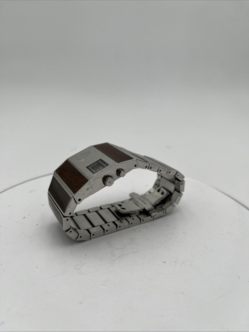May include: A silver wristwatch with a rectangular face and a stainless steel strap. The watch face has a dark brown wood-grain design and two small buttons. The watch is displayed on a white surface.