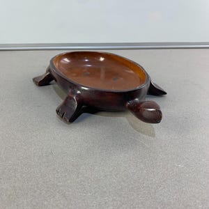 Può includere: Un piatto a forma di tartaruga in legno marrone scuro. Il piatto ha una ciotola poco profonda di forma ovale e quattro zampe. La tartaruga è scolpita da un unico pezzo di legno e ha una finitura liscia e lucida. Il piatto viene probabilmente utilizzato per contenere piccoli oggetti.
