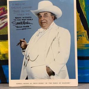 May include: A color photograph of Sorrell Booke as Boss Hogg from The Dukes of Hazzard. He wears a white cowboy hat, suit, and holds a cigar. The image includes handwritten text and a signature.