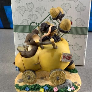 May include: A whimsical figurine featuring a mouse driving a yellow vehicle made from a gourd. The vehicle is loaded with acorns and berries. The base is decorated with flowers and grass, and the figurine is set against a box with leaf patterns.