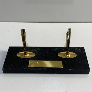 May include: A black marble pen holder with two gold pen stands. Each stand has a circular base and a vertical pen holder. A gold nameplate is centered on the base.