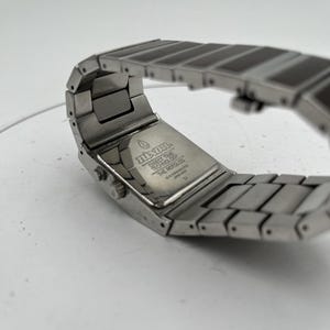 May include: A close-up of a silver wristwatch with a rectangular face and a linked band. The watch has a brushed metal finish and the text "Nixon" is visible on the back. The watch band is made of rectangular links.