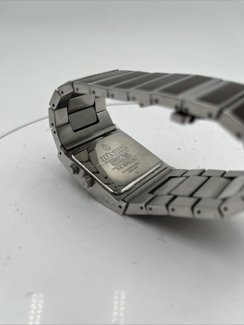 May include: A close-up of a silver wristwatch with a rectangular face and a linked band. The watch has a brushed metal finish and the text "Nixon" is visible on the back. The watch band is made of rectangular links.