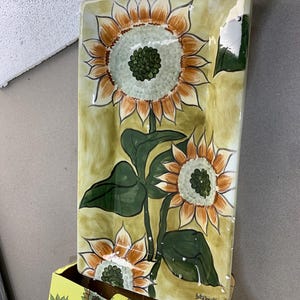 May include: Rectangular ceramic serving platter with a hand-painted sunflower design. The sunflowers have orange petals, white centers, and green leaves. The background is a light olive green. The platter is displayed with its original yellow box, also decorated with sunflowers.