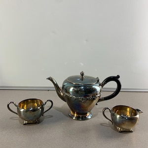 Peut inclure: Service à thé en argent comprenant une théière, un sucrier et un pot à crème. La théière a une poignée noire et un bord décoratif. Le sucrier et le pot à crème ont de petites poignées. L'intérieur des pièces est doré.