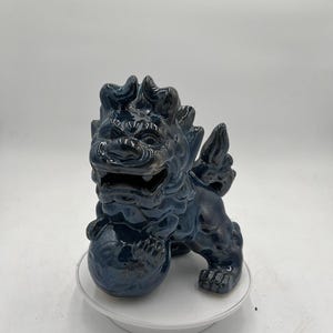 Puede incluir: Una estatua de león guardián de cerámica azul oscuro con un acabado brillante. La criatura mítica sostiene una bola y tiene detalles puntiagudos en la espalda y las alas. La estatua está sobre una superficie blanca.