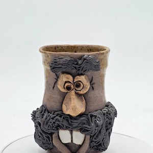 Könnte beinhalten: Eine handgefertigte Keramiktasse mit einem skurrilen Gesichtsdesign. Die Tasse hat einen braunen Boden mit einem beigen Rand und ein geformtes Gesicht mit großen Augen, einer Nase, einem Bart und zwei großen Zähnen. Die Tasse ist ein einzigartiges Kunstwerk.
