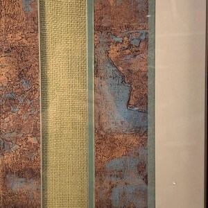 Puede incluir: Obra de arte abstracta enmarcada con paneles texturizados en tonos marr&oacute;n y azul, separados por una secci&oacute;n central de arpillera beige. La obra est&aacute; encerrada en un marco blanco, creando una est&eacute;tica moderna.