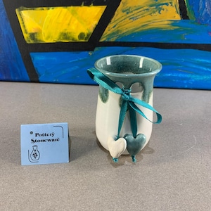 May include: A small, white ceramic vase with a teal rim and a teal ribbon tied around it. Two heart-shaped ornaments hang from the ribbon. A blue tag with the words "Pottery Stoneware" is on the left.