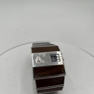 May include: A vintage digital watch with a silver metal strap and a rectangular face. The face has a dark wood-grain finish and a small digital display. The watch features the Nixon logo and a small digital display.