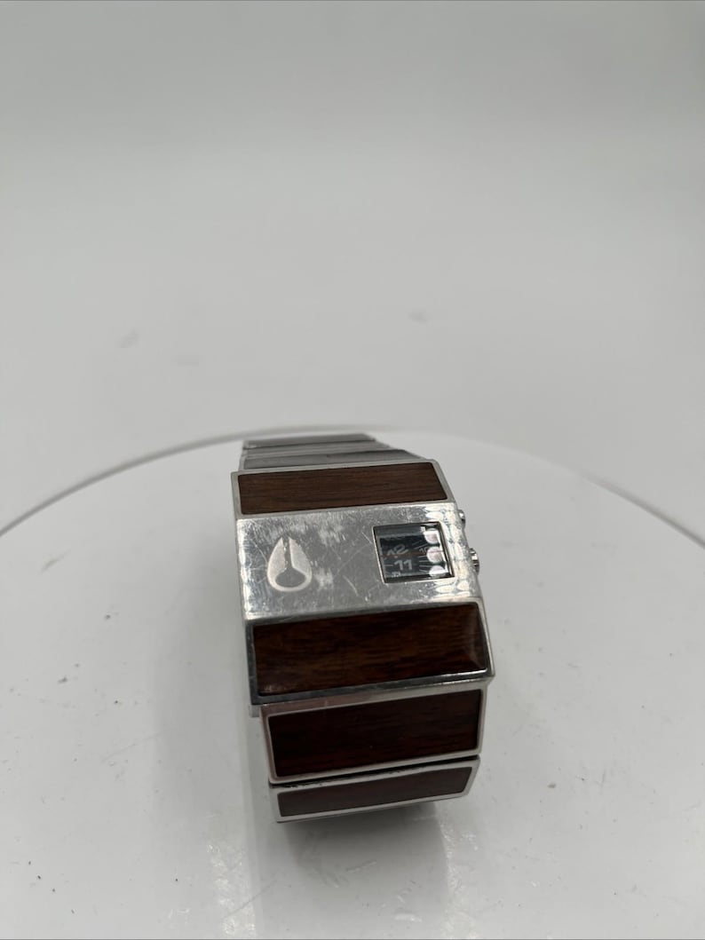 May include: A vintage digital watch with a silver metal strap and a rectangular face. The face has a dark wood-grain finish and a small digital display. The watch features the Nixon logo and a small digital display.
