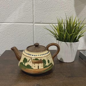 Puede incluir: Una tetera de cerámica con tapa, boquilla y asa marrones. El cuerpo es de color crema con una escena de paisaje pintada que representa una casa, árboles y la palabra "St Ives". La tetera está sobre una superficie de madera.