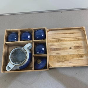 Peut inclure: Un coffret en bois contenant un service à thé en céramique bleu et blanc. Il comprend une théière avec une passoire, quatre petites tasses bleues et un couvercle bleu. L'ensemble est présenté dans une boîte en bois.