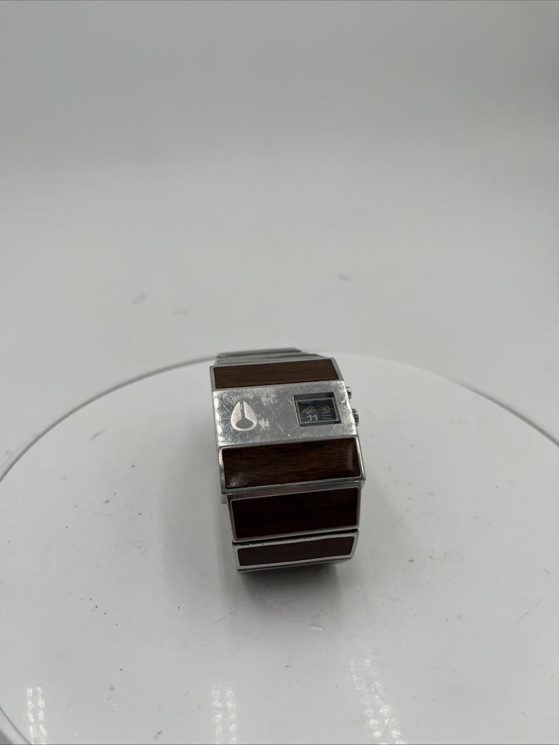 May include: A vintage Nixon digital watch with a rectangular face and a brown wooden strap. The watch features a silver bezel and a digital display. The Nixon logo is visible on the face.