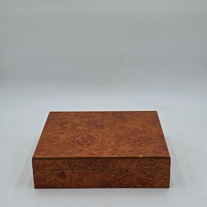 Puede incluir: Una caja de madera rectangular con un patrón de veta de madera de burl. La caja es de color marrón cálido y tiene una tapa con bisagras. La caja está cerrada y se encuentra sobre una superficie blanca.