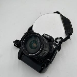 Op de afbeelding: Een zwarte Canon EOS D60 digitale camera met een bevestigde Sigma Compact Hyperzoom 28-300mm 1:3.5-6.3 lens. De camerabehuizing bevat het Canon-logo en de tekst EOS D60, terwijl de lens tekst bevat die aangeeft dat deze in Japan is gemaakt.