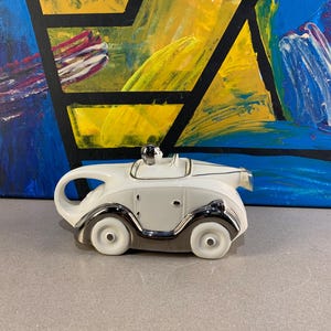 Peut inclure: Une théière en céramique blanche en forme de voiture ancienne, avec des détails argentés. La théière a une poignée incurvée, un petit bec et un couvercle avec un bouton argenté. Les roues et les garnitures sont argentées.