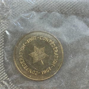 Op de afbeelding: Een goudkleurige munt met de tekst "CONFEDERATION" en de jaartallen "1867" en "1967", rondom een zespuntige ster. De munt is verpakt in een transparante plastic hoes.