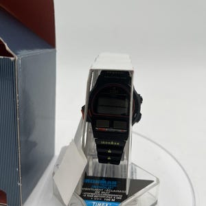 May include: Black Timex Ironman digital watch with red accents, displayed on a clear plastic stand. The watch features a black band and a digital display. The packaging includes the text "IRONMAN INDIGLO NIGHTLIGHT".