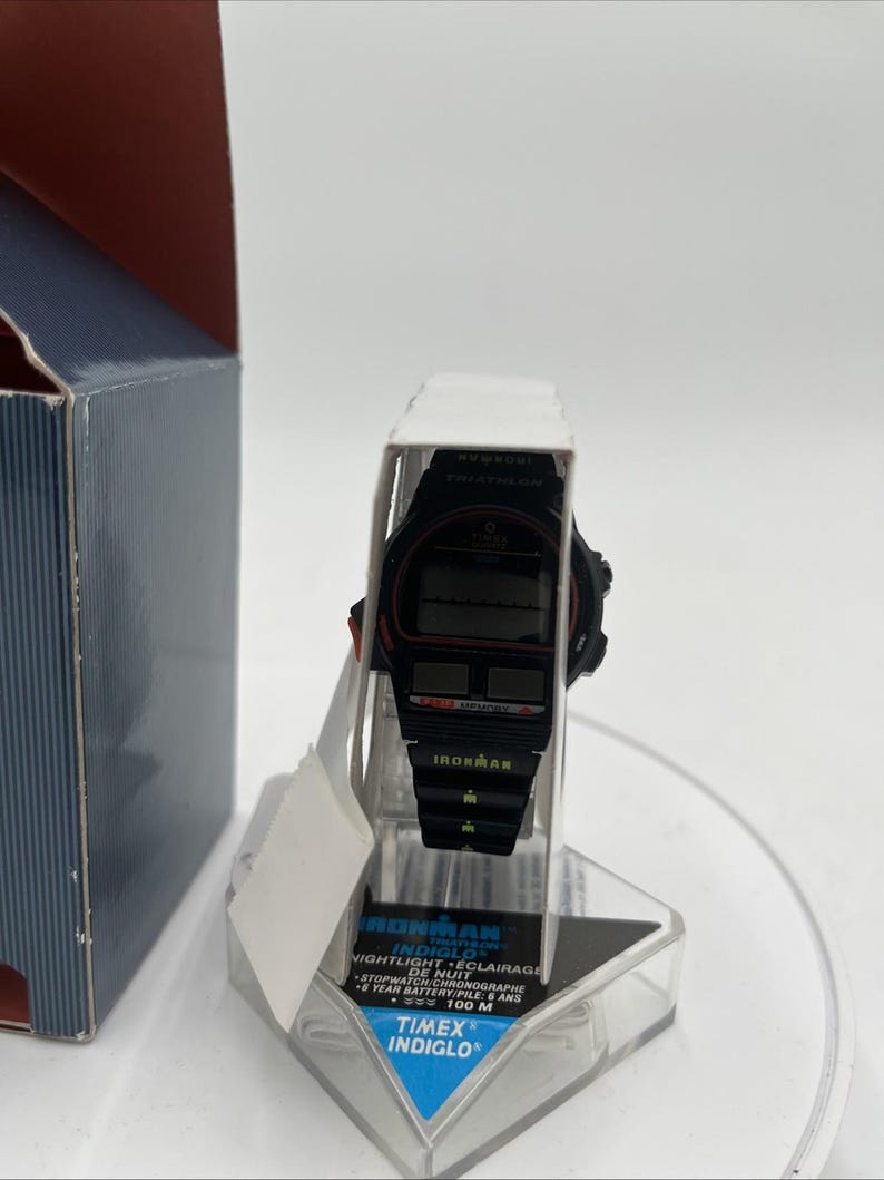 May include: Black Timex Ironman digital watch with red accents, displayed on a clear plastic stand. The watch features a black band and a digital display. The packaging includes the text "IRONMAN INDIGLO NIGHTLIGHT".