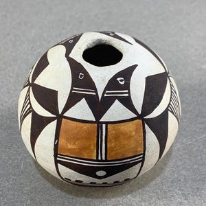 May include: A spherical ceramic pot with a small opening at the top. The off-white surface is adorned with black and brown geometric designs and bird motifs. A band of orange-brown colour is near the base, with a diameter of approximately 15 cm.