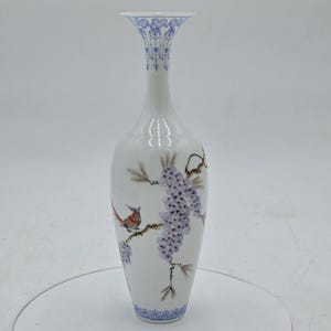 Può includere: Un vaso di ceramica bianco con collo sottile e bordo svasato. Il vaso presenta un disegno dipinto di un uccello appollaiato su un ramo con fiori di glicine viola. Motivi floreali blu adornano il bordo e la base. Il vaso è alto circa 25 cm.