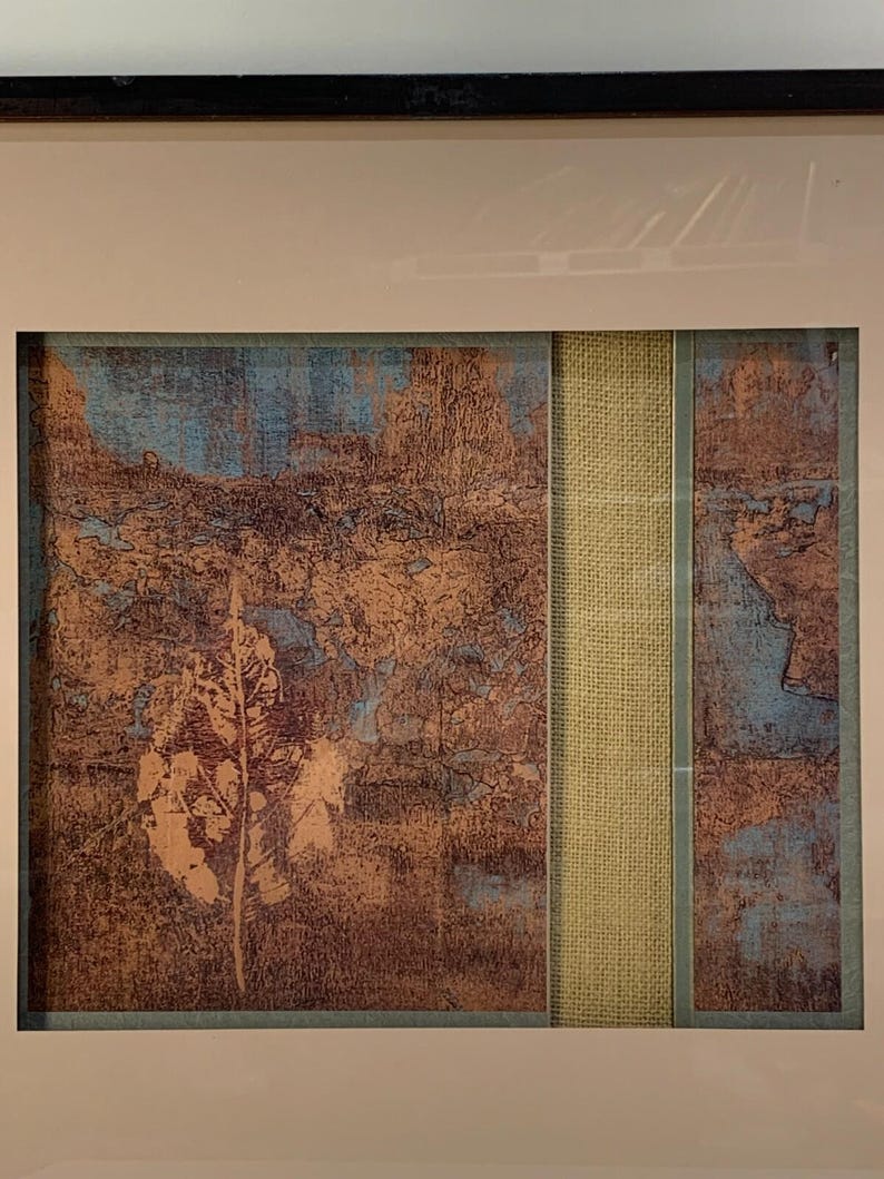Puede incluir: Obra de arte abstracta enmarcada con una apariencia texturizada. La obra presenta una combinaci&oacute;n de tonos marrones, azules y beige, con una tira vertical central de tela beige. El dise&ntilde;o general sugiere un tema de paisaje o natural.