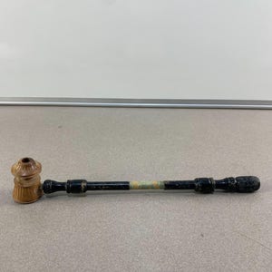 May include: A wooden pipe with a light brown bowl and a black stem. The stem has a decorative band of green and gold. The end of the stem has a textured design.