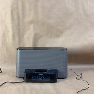 May include: A gray Sony clock radio with a digital display showing the time 11:25. The radio has a rectangular shape with a speaker grill on the front. A smaller black digital clock is in front of the radio.