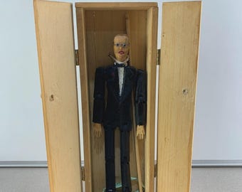 Puede incluir: Una marioneta de madera vintage en una caja de madera. La muñeca viste un traje negro con camisa blanca y pajarita. La caja tiene dos puertas con bisagras que se abren para revelar la muñeca. La muñeca tiene rasgos faciales pintados y está suspendida por cuerdas.