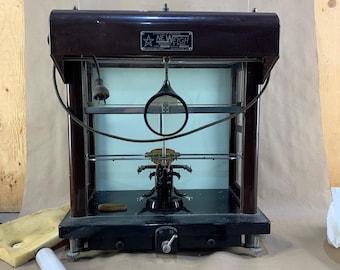 Puede incluir: Una antigua balanza New Weigh con una carcasa de vidrio y un marco marrón oscuro. La balanza tiene una base negra y una lente de visualización circular. Varios pesos y herramientas de medición están dispersos alrededor de la balanza.