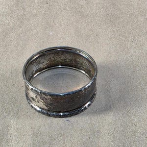 May include: A tarnished silver napkin ring with a textured surface. The ring is circular with a slightly raised, decorative edge. The metal shows signs of age and wear.