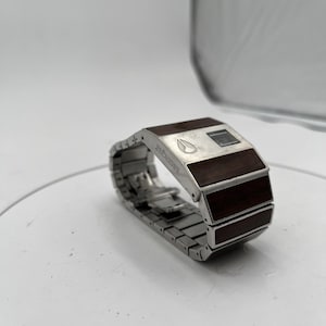May include: A silver wristwatch with a rectangular face and a metal link strap. The watch face has a dark wood grain design and a small digital display. The band is made of linked metal segments. The brand name "Nixon" is visible on the watch face.