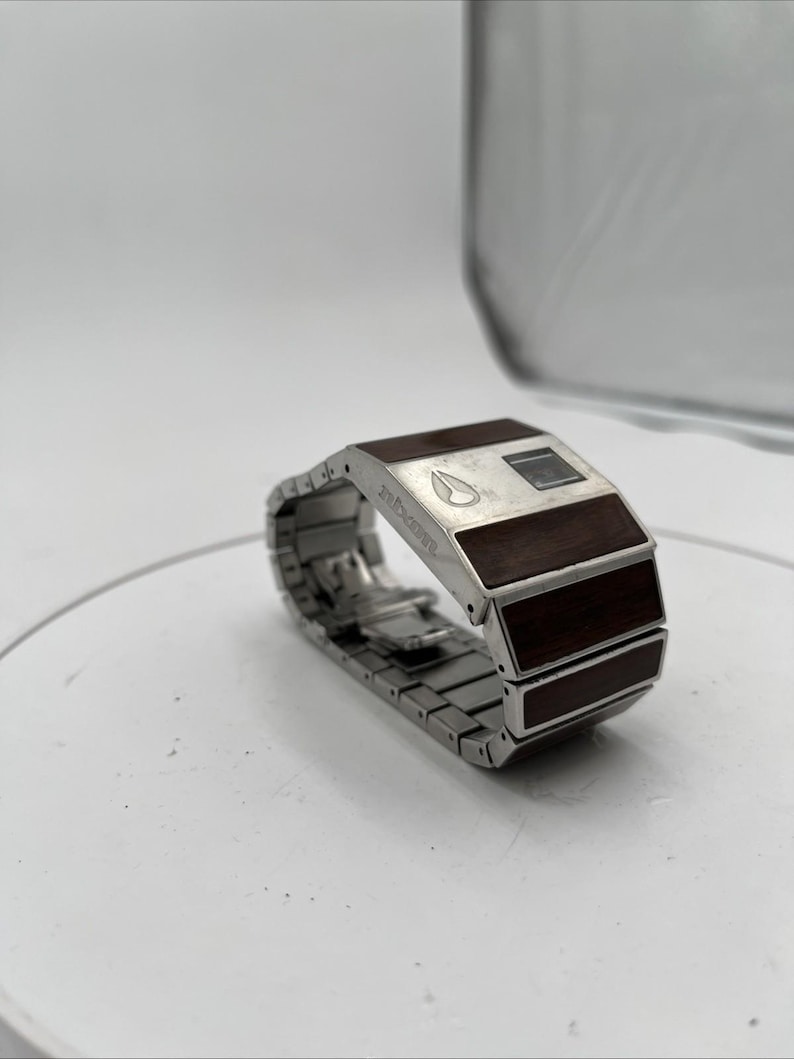 May include: A silver wristwatch with a rectangular face and a metal link strap. The watch face has a dark wood grain design and a small digital display. The band is made of linked metal segments. The brand name "Nixon" is visible on the watch face.